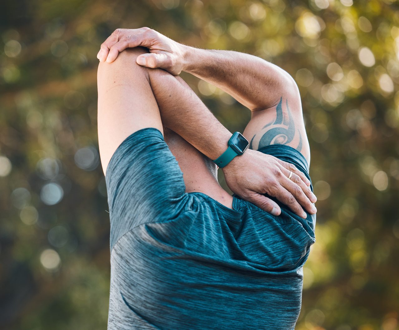 A man stretching his arm outdoors in a park with sports gear.